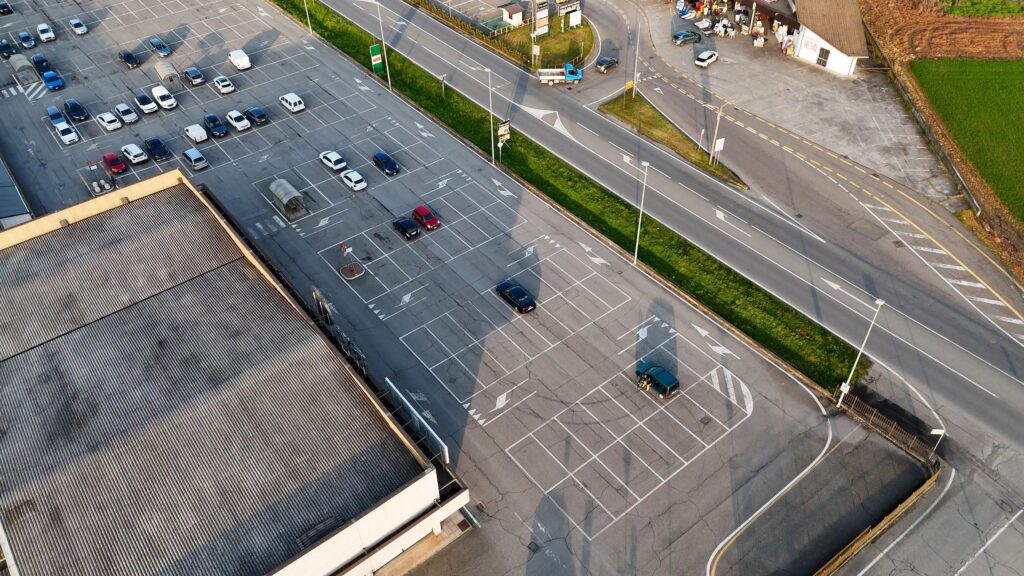 Aerial drone view of an extensive parking lot beside a commercial building and busy road in Castelvetro Piacentino, showing cars, road markings and green buffers