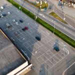 Aerial drone view of an extensive parking lot beside a commercial building and busy road in Castelvetro Piacentino, showing cars, road markings and green buffers